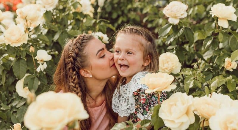 woman kissing girl's cheek between white roses garden